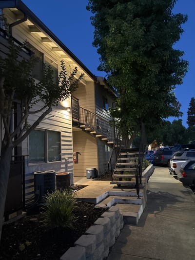 The image shows an exterior view of an apartment building at dusk. The building has light-colored horizontal siding and is illuminated by outdoor lighting. There is a set of metal stairs leading to an upper-level walkway. In front of the building, there are two air conditioning units, a small clump of ornamental grass, and a low retaining wall made of interlocking stones. A paved walkway runs alongside the building, and parked cars are visible in the background. The sky is a deep blue, indicating twilight. The scene is located in Sacramento, United States.