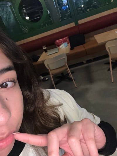The image shows a person's face, particularly their eye and mouth. The person's dark brown hair is visible, and they are wearing a white jacket with a black collar. Their right index finger touches their upper lip. A mole is visible near their nose. In the background, there are chairs and tables, suggesting a cafe or restaurant setting. The perspective is close-up, angled, and presumably a selfie.