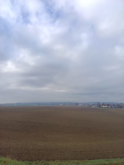 The image displays an expansive landscape identified as the rural periphery of Hoenheim, France. The upper two-thirds of the frame are occupied by a dense, uniformly grey overcast sky, with minor variations in luminosity and occasional faint glimpses of lighter blue. The immediate foreground, at the bottom of the frame, features a narrow strip of low green vegetation. This transitions into a vast, open field of dark brown, tilled earth, characterized by visible furrows and clods consistent with recent agricultural preparation. In the distant background, a horizontal band of dense, muted green-grey trees or vegetation partially obscures the horizon line. To the right of the background, a cluster of residential buildings, representative of a small town or suburban area, is visible. These structures predominantly feature light-colored facades and roofs in shades of reddish-brown or grey. No individuals, specific actions, or significant interactions are discernible within the static scene.