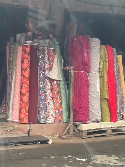 This is a medium-shot, eye-level photograph of a market stall in Tangerang, Indonesia, exhibiting an array of colorful fabric rolls. The stall is positioned in front of a building with an open facade, revealing stacks of white ceramic items behind the fabric. The fabric rolls, standing upright, display a variety of patterns and colors, including floral prints in pink, orange, and green on a brown background, abstract blue and white designs, and solid colors like burgundy, pink, green, yellow, and red. Some of the rolls are tied with white string. The stall has a wooden support structure and is situated on a raised platform. In the foreground, the ground is wet and appears to be a paved street or walkway, with some debris scattered on it. The overall lighting suggests an overcast day, with some glare visible on the fabric. The sign above the stall reads "BLOK A 5".
