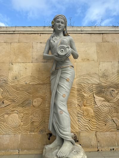 A full-length statue of a woman is affixed to a wall in Jimbaran, Indonesia. The statue is pale grey in color and depicts a woman lying on her side, holding a spherical object to her chest. She is adorned with a flower near her left ear, and gold-colored details are visible on her garment.
The wall behind the statue is made of tan-colored stone blocks, some of which are carved with depictions of fish and swirling water patterns. A narrow strip of blue sky and vegetation is visible above the wall.