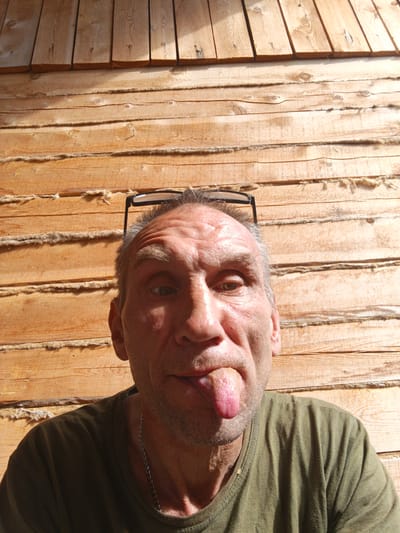A man with graying hair and glasses perched on his head sticks his tongue out at the camera. He is wearing a green t-shirt and a silver chain. The background is a wooden wall with horizontal planks. The image appears to be taken outdoors in Strelka, Russia.