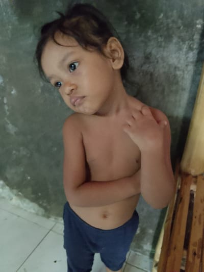 A young child with dark hair and brown eyes stands with their arms crossed, looking to the left. The child is shirtless and wearing dark blue pants. They are leaning against a textured, grey wall. To the right of the child, a section of a wooden structure with vertical planks is visible. The floor beneath the child appears to be tiled. The lighting suggests it is daytime. The child's gaze and posture might indicate contemplation or shyness. The setting appears to be an indoor or covered outdoor area, possibly a home or a communal space in Merbopuntong, Indonesia.