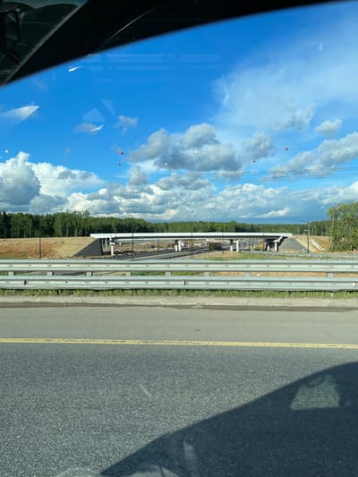 The image shows a view from inside a vehicle, possibly a car, driving on a road in Novodrozhzhino, Russia. The road has a double yellow line marking, a concrete curb, and what appear to be guardrails. Beyond the guardrails is a construction site with heavy equipment visible. The sky is blue with scattered white clouds. Reflections are visible on the vehicle's window. Overhead power lines with red and white markers are present. The image is rotated 90 degrees clockwise.