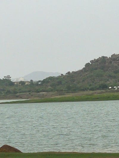 A landscape image presents a body of water, likely a lake or river, occupying the right portion of the frame. A lush green bank runs along the water's edge. A hill or low mountain, covered with dense vegetation, rises in the background. The sky is overcast, casting a diffused light over the scene. The location is near Nabatum, Nigeria.