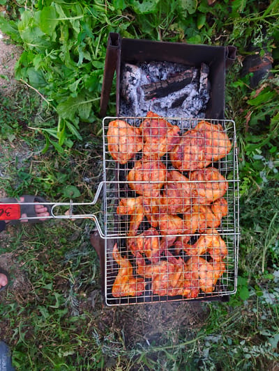 A close-up, overhead shot captures a grilling basket filled with golden-brown chicken pieces, likely drumsticks and thighs, arranged over glowing coals in a black metal barbecue. The basket is positioned within a rectangular grill, which sits on a grassy, outdoor surface. The surrounding environment is characterized by abundant green foliage, appearing somewhat wild and untamed, with scattered patches of dirt and pebbles. In the lower left corner, a person's foot, clad in a grey sandal, is visible, with a red-handled tool resting on the ground next to it. The overall scene suggests a casual outdoor meal being prepared in a rural or garden setting, possibly on the outskirts of Shchuchinsk, Kazakhstan, given the context provided.