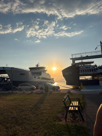In Keramoti, Greece, two ferries, "THASSOS I" and "THASSOS II," are docked at a harbor during sunset. The sky is filled with scattered clouds, illuminated by the setting sun, which casts a warm glow on the scene. In the foreground, a grassy area with a park bench is visible, and several cars are parked along the waterfront. The water is calm, reflecting the golden light of the sun. The overall impression is one of peacefulness and natural beauty.