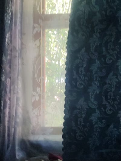 The image depicts a partially drawn window with curtains. On the left, a translucent, light purple curtain with a floral pattern is visible. To the right of this, a sheer white curtain with a more prominent floral design is present. The window itself, with its wooden frame, is visible behind the sheer curtain, and through it, lush green foliage and dappled sunlight can be seen, suggesting an outdoor environment. On the far right of the image, a dark blue, opaque curtain with an intricate, possibly damask or paisley, pattern is drawn closed, partially obscuring the view through the window. The bottom portion of the image is dark and out of focus, with a hint of fabric or objects on the floor. The scene appears to be indoors, looking out through a window. The mention of Smoline, Ukraine, in the prompt does not provide any visual cues within the image itself to confirm or deny this specific location.