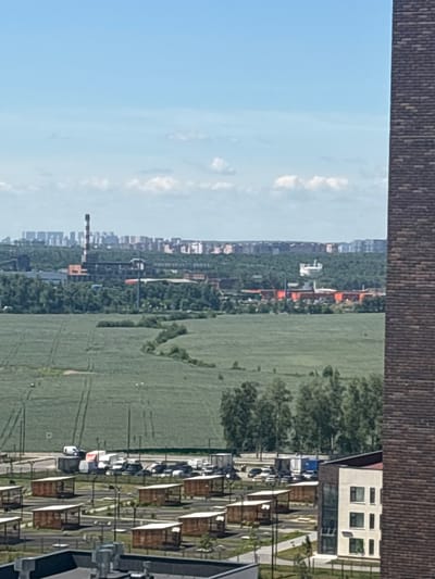 The image captures a panoramic view of Misaylovo, Russia, on a clear day with scattered white clouds. In the foreground, a brick building with a dark roof dominates the right side of the frame. Beyond it, a modern building with a white facade, multiple windows, and a dark roof is visible. Further into the scene, a parking lot filled with various vehicles, including cars, vans, and trucks, is organized near a row of small wooden structures with peaked roofs. A vast, green agricultural field stretches across the middle ground, marked by tire tracks. In the distance, a dense line of green trees separates the field from industrial buildings and a city skyline characterized by numerous residential high-rises and a prominent smokestack. The sky is a bright, clear blue.