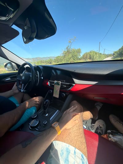 The image depicts the interior of a car with two individuals. The car appears to be in motion, as indicated by the perspective and the clarity of the scene. The dashboard features a red accent, a steering wheel, and a phone mounted with a navigation app displayed. One person is visible driving, with their hands on the steering wheel. Another person's legs and arm are visible in the passenger seat, and they are wearing a yellow watch. The exterior view through the windshield shows a sunny day with a clear blue sky and lush green vegetation, suggesting a rural or semi-rural environment. The road appears to be paved and winds through a hilly landscape. Based on the context of Arkadii Municipal Unit, Greece, the scene likely takes place on a road within that region, possibly en route to or from a destination in the area.