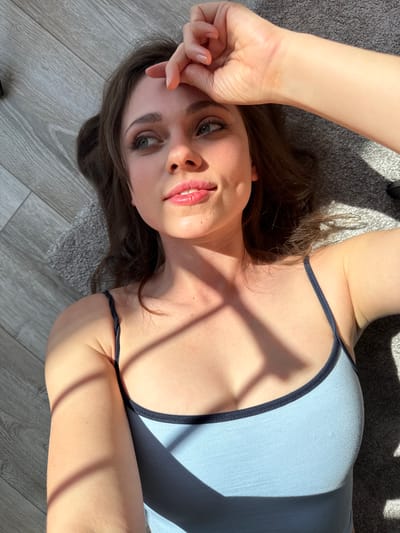 A woman is lying on a gray rug placed on a wooden floor. She is wearing a blue tank top with dark blue straps. Her right arm is raised, with her hand near her forehead. Sunlight and shadows play across her face and body. Her hair is brown and spread out on the rug. The location appears to be indoors and likely not in the city of Balatonlelle, Hungary as there are no recognizable landmarks or environmental clues to suggest that location.