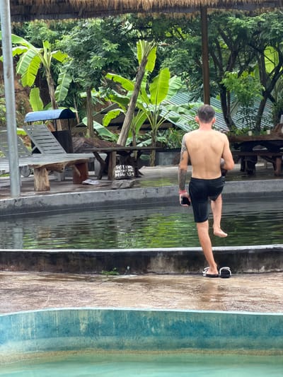 The image depicts an outdoor scene in Khanh Vinh, Vietnam, likely at a pool or water park. A shirtless man, wearing black shorts and flip-flops, is captured mid-stride as he walks along the edge of a pool. He is holding a mobile phone in his left hand and has tattoos on his left arm. The pool's water is a vibrant green, reflecting the lush vegetation surrounding the area, including banana trees and other dense foliage. In the background, there are wooden tables and chairs, as well as lounge chairs, suggesting a recreational setting. The ground around the pool is wet and appears to be a paved surface. A sign with Vietnamese text is partially visible on a wooden table in the middle ground. The overall environment is tropical and humid.