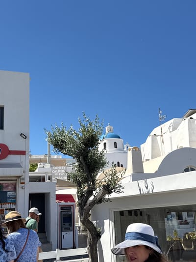 In Ia Municipal Unit, Greece, on a bright, sunny day, a scene unfolds on a street. A white building with a red stripe runs along the left side of the frame. To the right of this building, an olive tree with a gnarled trunk stands prominently. Beyond the tree, white buildings characteristic of Greek island architecture dominate the background, featuring domed roofs and arched windows. A prominent blue-domed church with a white cross stands out against the clear blue sky. In the foreground, several people are visible. A woman wearing a straw hat with a blue ribbon is partially in the frame on the right. To her left, a woman in a blue floral top and a straw hat is seen from behind. Further left, a man wearing a green shirt and a white baseball cap is talking on his phone, looking towards a building entrance with stairs. An ATM machine, branded "EUROBANK," is located to his right. Signs for "SUNSET CALDERA VIEW VOLCANO" are visible behind the olive tree, indicating the area's attractions. The overall impression is a bustling street scene in a picturesque Greek town.