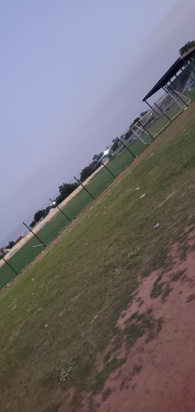 An outdoor, slightly tilted shot captures a sports field at dusk in Numan, Nigeria. The field, possibly a soccer pitch, has green turf and is bordered by a green fence. In the background, a covered seating area with a dark roof is visible, along with a white soccer goal. Trees line the edge of the field. The sky is a pale blue, indicating the end of daylight. Scattered on the grass are a few white plastic bottles, suggesting recent activity or litter. A path or dirt area runs along the bottom right of the frame. The image has a somewhat grainy quality, consistent with low-light photography. There are no people clearly visible in the frame.