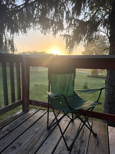 This image shows a green folding chair on a wooden deck. In the background, there is a sunrise over a grassy field with trees. The sun is low on the horizon, casting a warm glow over the scene. The deck appears to be weathered, with visible wood grain and nail heads. A tree trunk is visible on the right side of the image, and tree branches hang down from the top. The overall impression is one of a peaceful morning setting, likely in a rural or suburban area. The location is identified as the city of Holt, United States.
