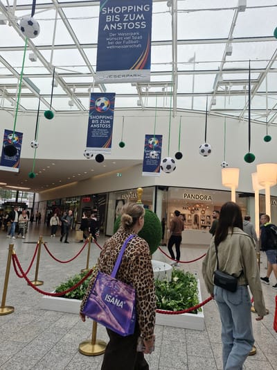 The image depicts an indoor shopping mall in Bremen, Germany, likely decorated for a football event. Suspended from the ceiling are several footballs and decorative green balls, along with banners that read "SHOPPING BIS ZUM ANSTOSS" (Shopping Until Kick-off) and "WESERPARK" (Weser Park), a shopping center located in Bremen. The mall's interior features a tiled floor, a high ceiling with a visible glass roof, and store fronts. In the foreground, two women are walking away from the camera. The woman on the left is wearing a leopard-print jacket and carrying a purple shopping bag with "ISANA" written on it. The woman on the right is wearing a beige jacket and blue jeans, with a black crossbody bag. Other shoppers are visible in the background, as are display lamps and a large green spherical decoration. A red rope barrier with gold stanchions is also present in the foreground, sectioning off a decorative planter.