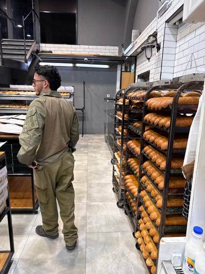 A man in a khaki shirt and pants stands in a bakery located in the city of Kiryat Ekron, Israel. The man is facing a rack of baked bread. He has short, dark hair and is looking at the bread. The bakery has white tiled walls and a wooden floor. There is a metal staircase in the background. The bread is arranged in metal racks on wheeled metal bases. The bread is all similar, with a light brown crust and a slightly darker inside.  The bread is stacked and arranged in rows. The bakery is well-lit and clean.