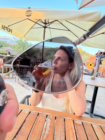 In Zagora, Montenegro, a woman is reflected in sunglasses while holding a glass of beer. She is wearing a white tank top and has blonde hair. The background shows an outdoor seating area with tables and chairs, as well as mountains and trees under a clear blue sky. A Pepsi umbrella is visible in the upper portion of the image.