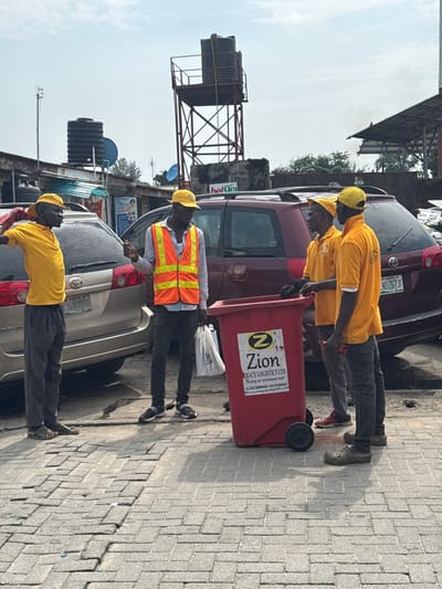 This image depicts an outdoor urban scene in Ajah, Nigeria, under a bright, slightly overcast sky. Four adult males are present on a paved surface made of interlocking concrete blocks.
A red wheeled waste bin, approximately waist-height, is centrally located in the foreground, branded with "Zion GRACE LOGISTICS LTD." and the slogan "Keeping our environment clean," along with contact details.
Three of the men are positioned around this bin, appearing to interact. The central figure wears a light blue long-sleeved shirt, an orange reflective safety vest, dark jeans, and a yellow baseball cap; he is gesturing with his right hand while facing right. To his right stand two men, both in yellow short-sleeved shirts (with visible logos), yellow baseball caps, and grey pants. The man closer to the center holds dark gloves; the man furthest right holds a red object in his right hand. Both face left, towards the man in the safety vest. The fourth man is positioned to the far left, wearing a yellow polo shirt, a yellow baseball cap, grey baggy trousers, and dark sandals, with his left arm partially raised.
Behind the individuals, two minivans are parked: a silver Toyota minivan (partially visible, with a license plate "LAGOS ND-206 JL" or similar) and a dark red minivan (with license plate "LAGOS NGN0267 JF"). The background reveals a commercial or mixed-use area featuring various structures, including buildings with metal roofs, partial signage (e.g., "hotQia," "Shop With Us"), and two large black plastic water tanks. One tank is elevated on a tall, red-painted metal gantry, while another is on a lower, less prominent structure.