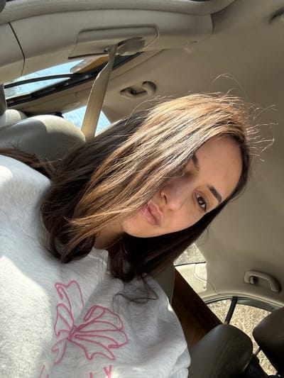 The image displays a female individual situated inside a vehicle. Her medium-length, dark brown hair partially covers the left side of her face. Her visible right eye is dark, as are her eyebrows. Her lips are full and slightly pouted. She wears a light grey crewneck sweatshirt with a large pink embroidered bow design on the chest. The vehicle's interior features light tan or beige upholstery on the seats and headliner. A beige seatbelt is visible, extending downwards from the ceiling on the upper left. Through a partially open sunroof on the upper left, a clear blue sky and sparse tree branches are discernible. A beige grab handle is present on the right side of the vehicle's ceiling. The individual is oriented towards the camera. The specific city of Golyamo Belovo, Bulgaria, cannot be determined from the visual information within the image.