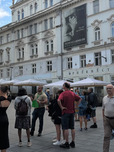 The image depicts a street scene in Sarajevo, Bosnia and Herzegovina, focused on a building facade displaying a large banner for the "Srebrenica Genocide Memorial Exhibition." Several people are visible in the foreground, standing on a paved area.
The building is multi-story with ornate architectural details. A prominent banner on the upper portion of the building features a black and white close-up of an eye and text that reads "You are my witness" and "SREBRENICA GENOCIDE MEMORIAL EXHIBITION." The date "GALLERY 11/07/95" is also present. Below the banner, the word "PHARMACY" is visible in large letters above a street-level entrance. White umbrellas, likely from outdoor seating for a cafe or restaurant, are positioned in front of the building.
In the foreground, a group of individuals are gathered. On the far left, a woman in a black dress is partially visible. Next to her, a woman with her back to the camera wears a grey hoodie and a patterned skirt, with a backpack. A man in a tan t-shirt and jeans is speaking, holding a green bag. To his right, a woman with blonde hair in a denim jacket is looking at her phone. Further to the right, a man in a red t-shirt and shorts stands with his hands on his hips. Another man in a dark jacket and shorts is also present, as is a man in a light-colored polo shirt and brown pants on the far right. The ground is paved with square tiles. The overall lighting suggests an overcast or partly cloudy day.