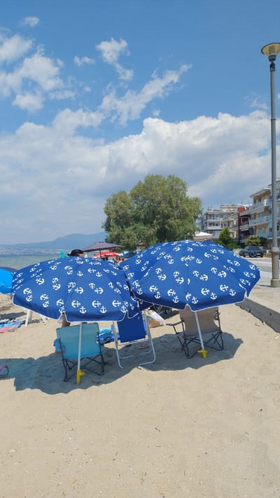 This image depicts a section of a sandy beach in Neoi Epivates, Greece, under a bright blue sky with scattered white clouds. Two large blue beach umbrellas with white anchor patterns are prominently displayed, providing shade for beach chairs. One umbrella is positioned to the left, and another is to its right, slightly overlapping.

Beneath the umbrellas, several beach chairs are visible. To the left, a light blue chair with a metal frame is placed. Next to it, a darker blue chair. To the right of the second umbrella, a beige folding chair with a metal frame and black fabric is set up. A person's legs are visible through the gap in the right umbrella, and another person's feet are visible in the vicinity of the beige chair. A picnic basket is also tucked away under the right umbrella.

In the background, a line of trees with dense green foliage stands beyond the beach. Further back, buildings with balconies and multiple floors are visible, suggesting a coastal town or resort area. A street lamp stands tall to the right of the frame. The sea is visible in the distance, with a mountain range forming the horizon line. The sand on the beach appears fine and light brown, with some small shells and debris scattered across it. The overall scene captures a typical summer day at a Greek beach.