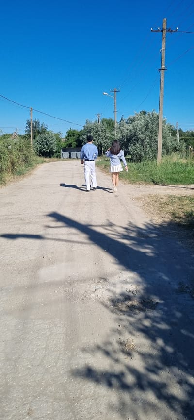 In Blizhnee, Ukraine, a man and a woman stand on a dirt road under a clear blue sky. The man, wearing a light blue button-down shirt and white pants, faces away from the camera, his shadow stretching across the road. The woman, in a light blue long-sleeved shirt and a white pleated skirt, also faces away from the camera and holds a white object in her hand. She is positioned slightly ahead of the man.
The scene is rural, with trees and tall grass lining the sides of the road. A telephone pole with multiple wires stands to the left of the man, and another pole with a light fixture is visible further down the road. The sun casts sharp shadows, suggesting it is midday. The overall impression is one of a casual outing on a bright, sunny day. No text is visible in the image.