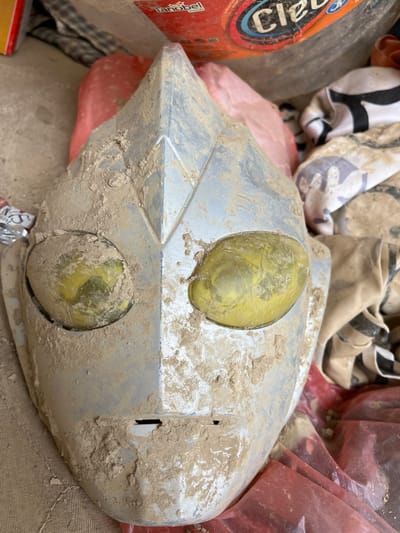 A close-up shot shows a silver, futuristic-shaped object, possibly a car headlight, covered in dirt. Two yellow-tinted circular lenses are visible on its surface, also partially obscured by the dirt. The object is nestled among a pile of other items, including red plastic bags, what appears to be clothing with black and white patterns, and a partially visible orange container with blue lettering.

The overall scene suggests a chaotic or perhaps a debris-filled environment, possibly after an event like a flood or natural disaster, given the mud and scattered items. The lighting is neutral, offering no clear indication of the time of day or specific weather conditions beyond the presence of dirt. The location context of Kota Kuala Simpang, Indonesia, hints at a possible Indonesian setting.

The orange container has visible text, with "Cl" clearly legible in large blue letters, and "Tanobel FOOD" partially visible below it. Other indistinct markings and logos are also present on the container. The image focuses primarily on the dirt-covered object, emphasizing its texture and the way it is integrated with the surrounding debris. There are no people visible in the frame. The activity suggested is one of aftermath or storage of recovered items, rather than an active event.