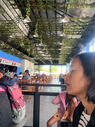 The image depicts an indoor dining area with a high ceiling adorned with artificial greenery and industrial-style pendant lights. The overall ambiance suggests a modern, casual restaurant or cafe. In the foreground, a woman is captured in profile, facing to the right with her eyes closed. Her dark hair frames her face, and she is wearing a dark jacket over a striped shirt. A portion of a red and grey backpack is visible on the left side of the frame, suggesting it belongs to someone sitting next to her.

In the midground, patrons are seated at wooden tables and chairs. A prominent blue wall with text reading "GACOAN IS READY" is visible on the left, indicating a food establishment. Further into the background, through large windows, glimpses of outdoor foliage and trees can be seen, suggesting the location is near a natural or green area. The restaurant appears to be moderately busy, with several tables occupied. The scene implies a relaxed atmosphere, possibly a popular spot in the city of Buleleng, Indonesia, known for its casual dining experiences.