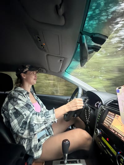 The image shows a woman in the driver's seat of a car, her body angled towards the passenger side. She is wearing a pink top and an open plaid shirt over it, along with a camouflage-patterned baseball cap. The car's interior is visible, including the steering wheel, dashboard with a navigation screen displaying a map, and a gear shift. The woman's left hand, wearing a ring, is on the steering wheel. Her legs are also visible.

The scene outside the car, viewed through the windshield and side window, appears to be a wooded area with tall trees and a blurred green and brown landscape, suggesting movement. The lighting suggests it is daytime. The mood appears relaxed, with the woman looking upwards, perhaps out the window or at something above. The car's interior lights are on, indicated by a small orange light on a panel near the ceiling.

The navigation screen displays text in Russian, including what appears to be a date and time reading "20:20.0". Other text on the screen includes "Bluetooth 203" and "2:46". The image is oriented sideways.