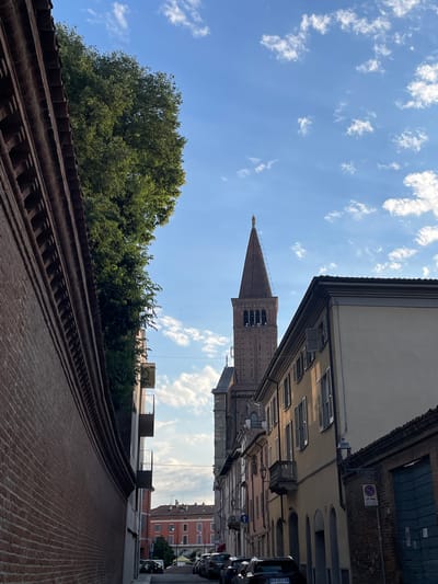 The image depicts a street view in Piacenza, Italy, oriented vertically. A tall, thin, brick tower with a pointed roof, potentially a campanile, rises between buildings. The sky is a bright blue with scattered white clouds. Buildings line the street, varying in height and color. A row of cars is parked along the right side of the street. In the foreground, there is a red brick wall, possibly part of a building or garden, with a tiled roof. A dense green tree is visible behind the wall. The architectural style suggests a historical European city center.