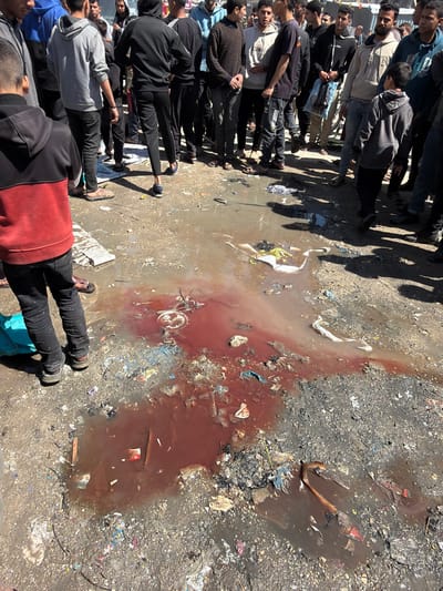 The image portrays an outdoor scene in Jabalia, Palestinian Territory, centered on an unpaved, uneven dirt and mud ground heavily littered with refuse. A significant, irregular-shaped puddle of reddish-brown liquid, consistent with blood mixed with water, dominates the foreground and midground. Within this liquid and surrounding debris, various items of trash are visible, including plastic remnants, fabric scraps, and what appears to be a white, circular object partially submerged in the most concentrated reddish area.
Approximately 20-30 males, including adults and several younger individuals, are gathered around the perimeter of the liquid, observing the scene. They are dressed in casual clothing, such as hooded sweatshirts, jackets, and pants. Many are looking downwards towards the ground. In the immediate foreground, a boy wearing a black top with red sleeves and black pants stands with his back to the viewer, facing the puddle. Shadows cast by the individuals suggest bright, direct sunlight. The background shows additional people and indistinct structures, indicating a populated area.