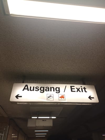 The image shows a white, illuminated sign with black text that reads "Ausgang / Exit". Arrows pointing left and right are also present on the sign. Below the text, there are two pictograms: one of a winged horse and the other of a person. The sign is mounted on a textured grey wall. To the left of the sign, a bright, vertical light source illuminates the scene. In the background, several other illuminated lights are visible along the wall, suggesting an indoor or covered public space, possibly a tunnel or underpass, given the lighting and wall texture. The setting appears to be dimly lit apart from the bright lights, implying it might be evening or night, or the area is underground. The location is indicated as Dortmund, Germany, which might explain the German text on the sign. There are no people visible in the image. The overall mood is utilitarian and informative, typical of signage in public transportation hubs or large buildings.