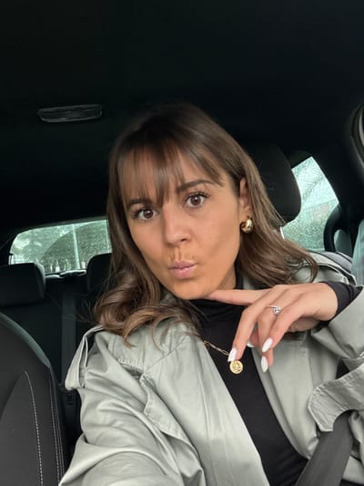 The image depicts an adult female situated within the passenger compartment of a vehicle in Villeneuve-Loubet, France. The vehicle's interior is dark, featuring black seating with visible white stitching and a black overhead liner. The female subject has brown hair with bangs, brown eyes, and directly faces the camera. Her lips are pursed, and her left index finger is placed beneath her chin. She is dressed in a black high-necked garment underneath a light gray trench coat. Accessories include gold-colored hoop earrings, a gold-toned necklace featuring a circular pendant with the Libra zodiac symbol, and a ring with a clear stone on her left ring finger. Her fingernails are painted white. A seatbelt is engaged across her chest. Through the vehicle's rear window, blurred green foliage and precipitation on the glass are discernible, suggesting outdoor conditions. No specific landmarks or precise street location within Villeneuve-Loubet are identifiable from the image.