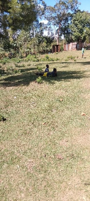 The image shows a grassy field with two individuals lying down in the distance. One person is lying on their back with their legs extended and the other person is lying on their side, facing the first person. Both are wearing dark colored clothing. There are trees and foliage surrounding the field and a few structures visible in the background. This image appears to be taken in a rural area outside the city of Bungoma, Kenya, at the coordinates 0.554 latitude, 34.773 longitude.  The light in the image suggests the time of day is midday or early afternoon.