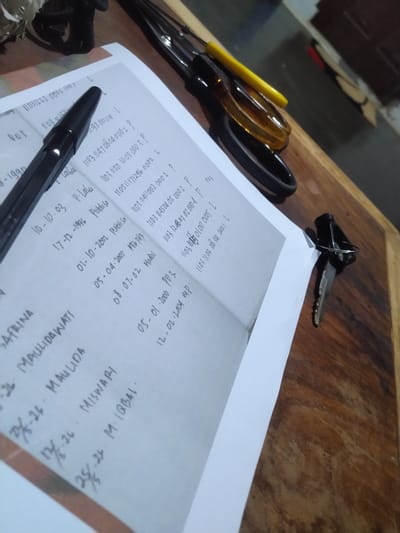 An angled, close-up shot captures an array of objects on a wooden table or desk. The scene is illuminated, suggesting a daytime or well-lit indoor setting.

In the foreground, a black pen rests on a white sheet of paper that is covered with handwritten text. To the left of the paper, a pair of screwdrivers, one with a yellow handle and another with an amber-colored handle, are visible alongside a black ring or hair tie. A set of keys, featuring a silver key attached to a black keyring, is positioned above the paper on the wooden surface. The background is blurred, showing a dark floor and some indistinct objects or furniture.

The paper displays extensive handwritten information, including names, dates, and numerical codes. Recognizable names include "SAFRINA," "MAULIDAWATI," "MAULIDA," "MISWARI," and "M. IQBAL," often associated with fractions like "25/26" or "17/26." Various dates such as "10-10-03," "17-12-1996," "01-10-2002," "05-04-2005," "08-07-02," "05-01-2000," and "12-02-2004" are present. Other words like "P.Labu," "Putonsa," "MT6 Pdg," "Madal," "MP," and "ROT" can be discerned. Numerous numerical sequences, frequently starting with "1103," are written in columns, potentially representing identification or record numbers, with some lines ending in "P." or "L.". The arrangement of the pen and paper suggests an activity of writing, record-keeping, or studying. The presence of tools and keys indicates a practical workspace or a personal area within Gampongbaro, Indonesia.