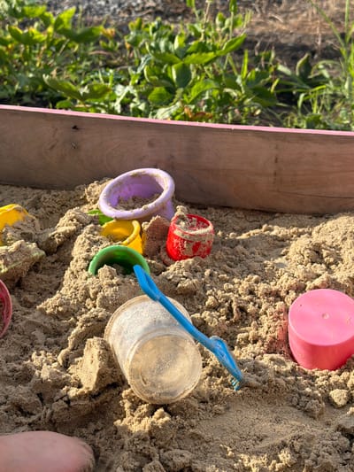 The image depicts a close-up view of a sandbox filled with sand. Several children's toys are scattered within the sand, including a pink plastic cup, a small red plastic cup, a purple ring-shaped toy, a yellow bucket, a green spade, and a clear plastic container. A blue plastic shovel is also visible, partially buried in the sand. The corner of a wooden sandbox structure with a pink border is visible on the left side of the frame, along with some green foliage and dry grass. A person's foot, covered in sand, is visible in the bottom right corner of the image. The lighting suggests it is daytime, likely sunny. The overall scene suggests a recreational activity, possibly children playing in a sandbox. There is no visible text in the image. The location context of Krasnyi Krym, Russia is noted.