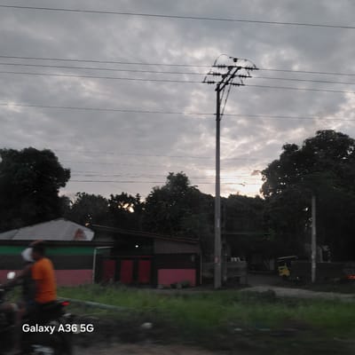 The image depicts an outdoor scene with a cloudy sky and a pole with electrical wires dominating the left side. In the background, there are trees and some buildings with red and green accents. The sun is visible through the clouds, suggesting it is either sunrise or sunset. On the right side of the image, a person on a motorcycle is visible, wearing an orange shirt and a white mask. The text "Galaxy A36 5G" is overlaid on the lower right corner of the image, indicating the device used to capture the photo. The scene appears to be in Carmen, Philippines, as suggested by the location context. The overall mood is somewhat overcast, with the warm glow of the setting or rising sun offering a contrasting element. The presence of electrical infrastructure and simple buildings points towards a residential or semi-rural area.