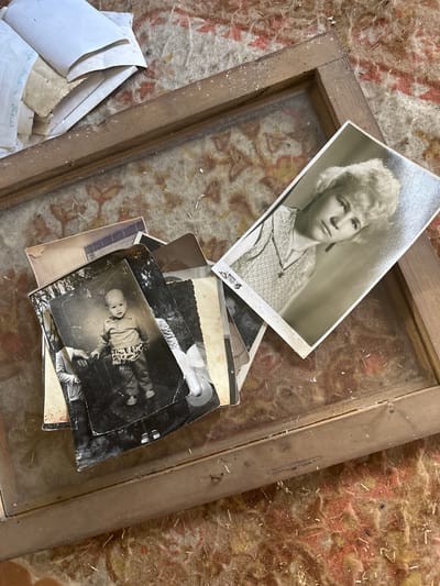 The image shows a collection of old black and white photographs resting within a weathered wooden frame on a textured surface. The photographs depict individuals, including a young woman with short blonde hair and a baby in a patterned outfit. Some loose papers are visible near the frame, and a small portion of handwritten Cyrillic script can be seen on one of them. The background is out of focus but suggests a rough, textured surface. The location is Студзёнкаўскі сельскі Савет, Belarus.