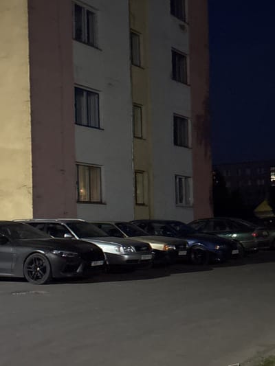 The image captures a nighttime scene in Лабжанскі сельскі Савет, Belarus, featuring a line of vehicles parked on an asphalt surface in front of a multi-story residential building.
The building, illuminated dimly, displays a facade composed of distinct vertical sections: a pale yellow/beige on the far left, a central light pink/mauve column, and a predominant white/light grey section. Multiple dark-framed windows are visible across its upper floors. On the lower-middle right, one window emits a warm, orange-toned light, revealing a blurry, indistinct red object within.
Five distinct vehicles are visible in the foreground, arranged in a queue from left to right:
1. A dark grey or black modern sports coupe with a low-slung profile, aggressive front fascia, and multi-spoke black wheels. Its front license plate displays the Belarusian alphanumeric sequence "3805 IX-S".
2. Immediately to its right, a silver sedan, likely an older model from the late 1990s or early 2000s, is partially visible. A partial Belarusian license plate "3331 BI-" is discernible on its front.
3. Next, a dark blue sedan, mostly obscured by the silver vehicle, shows minimal detail.
4. Further right, a dark green sedan is partially visible, largely obscured by the blue vehicle.
5. Additional vehicles are dimly perceived further down the parking row, their details obscured by darkness.
The ground is a dark asphalt lot. The sky is dark blue/black, indicative of nighttime. No individuals or specific actions are observed within the frame. In the far background, a darker building outline and some foliage are faintly visible. A yellow conical object is present on the far right edge of the image.