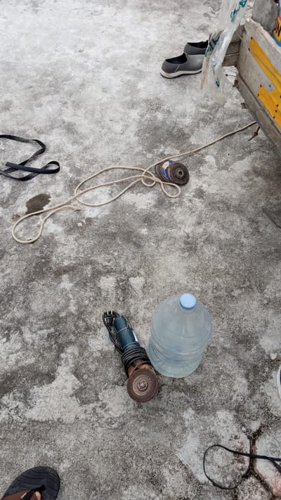 A high-angle medium shot captures an outdoor scene on a rough, concrete surface. In the upper right quadrant, a pair of grey slip-on shoes are placed next to a wooden structure with yellow accents and a clear plastic bag. Below and to the left of the shoes, a coiled rope lies on the concrete, with a small stack of metal discs positioned near its center. To the left of the rope, a black, flat strap is also present. In the lower half of the image, a small, blue-handled grinder with a metal cutting disc is situated next to a large, clear plastic water jug with a blue cap. In the bottom left corner, a person's left foot wearing a black flip-flop is visible. The scene appears to be on a ground level surface in Bireuen, Indonesia, potentially a workshop or outdoor storage area.