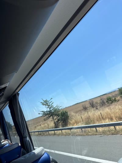 The image depicts a view from inside a vehicle, likely a bus, traveling on a road. The perspective is from a window looking outwards. In the foreground, part of the vehicle's interior is visible, including blue and gray seats. Gray curtains hang to the left.
The window frames a landscape characterized by dry, golden fields and sparse vegetation, suggesting a rural or semi-arid environment. A single, thin tree stands in the middle ground, with a few scattered bushes. A metal guardrail runs along the side of the road. The sky is a clear, bright blue, indicating a sunny day. In the far distance, a faint line of hills or mountains is visible.
While the image itself does not contain specific markers of Belozem, Bulgaria, this scene is consistent with the typical rural landscapes found in the region, particularly during drier seasons. The scene is static, with no apparent activity or interactions taking place. The lighting suggests daytime.