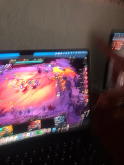 The image shows a laptop screen displaying a game, possibly a multiplayer online battle arena (MOBA) game. The game interface is visible, showing character icons and in-game action. The laptop is positioned on a surface, possibly a desk or table. Part of the keyboard is also visible. A second device is vaguely seen in the background, potentially a phone or tablet, showing a blurred screen. There is no evidence to suggest the location is in the city of Beccar, Argentina.