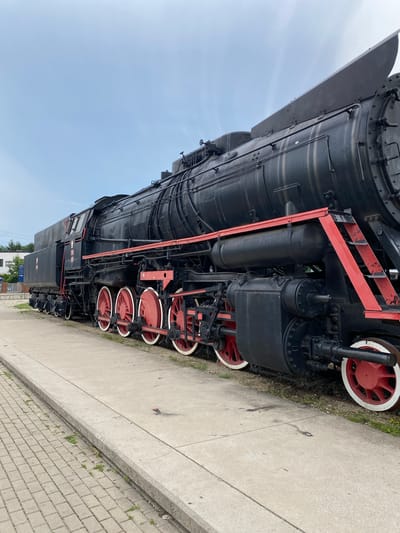 The image shows a large, black steam locomotive with red accents parked on a railway track alongside a concrete platform. The locomotive is oriented from left to right, with its front end not fully visible. Multiple large, red-spoked wheels with white rims are prominent along the side of the train. The sky above is a pale blue with some clouds. To the left of the train, a paved walkway made of interlocking bricks is visible, leading towards a building with some signage. The setting appears to be an outdoor railway station or museum. This steam locomotive is located at the Poznań Railway Museum (Muzeum Kolejnictwa w Poznaniu), part of the National Museum in Poznań, which is situated in the Wola district of Poznań, Poland.