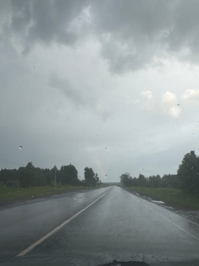 The image captures a view from inside a vehicle, looking out onto a wet road during what appears to be rainy or recently rainy weather. The perspective is through a windshield or window, as numerous clear raindrops are visible across the frame.

The central subject is an asphalt road, slick with water, extending into the distance under an overcast sky. A distinct white line runs along what appears to be the right edge of the visible road, guiding the eye towards the vanishing point. On both sides of the road, dense green foliage and trees line the horizon, suggesting a rural or wooded environment.

The sky is heavily clouded and gray, with varying shades indicating thick cloud cover and potentially ongoing precipitation. The dim lighting suggests either early morning, late afternoon, or simply a very gloomy day due to the weather. The overall scene conveys a sense of travel through a quiet, natural landscape under somber weather conditions.

No specific activity beyond "traveling" is explicitly shown, but the context implies driving. There is no visible text in the image. The location context for this scene is Otchor, Russia.