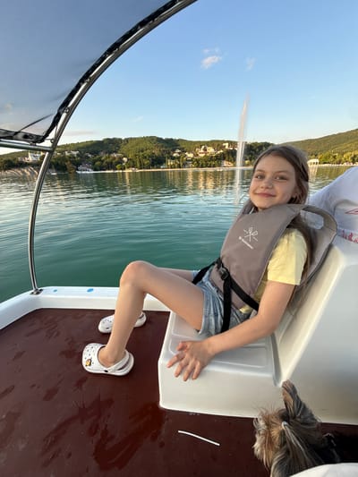A young girl with light brown hair, wearing a yellow t-shirt and denim shorts, sits on a boat with a life vest on. She is smiling at the camera. To her right, a small dog with long, dark fur is partially visible. The boat is on a body of water, with a green, tree-covered hill and buildings in the background. A large fountain sprays water into the air in the distance. The sky is clear and blue, suggesting it is daytime. The overall mood is happy and relaxed. A logo on the girl's life vest appears to read "BUOYABRAU."