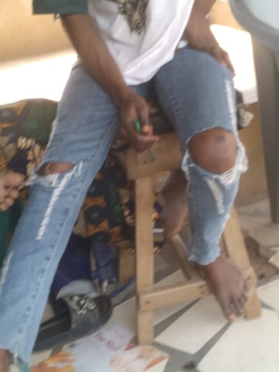 The image displays an individual seated in an indoor or semi-outdoor location within Agege, Nigeria. The subject wears a white t-shirt and light blue distressed jeans with prominent tears at both knees and along the upper thighs, exposing skin. The subject's forearms and lower legs are visible, displaying dark skin. The left hand grasps a small, slender object, green with an orange tip. The individual is barefoot, with a black slide sandal featuring white details resting on the tiled floor near the left foot. The person is positioned on a low, light-colored wooden stool. To the left, a multi-colored patterned fabric is visible. A dark blue object is partially obscured beneath the stool. The floor is covered with cream-colored tiles accented by darker patterned lines. A partially visible publication, "The Nail," lies on the floor in the foreground.