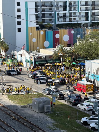 This is an elevated, eye-level, full shot of a street scene in Hollywood, Florida, USA. A large crowd of people, many wearing yellow, are gathered on the sidewalk and street. Several police cars are present, some with their lights on, and an ambulance is visible. Buildings line the street, including a multi-story apartment building with balconies and a building with a colorful, striped facade. Traffic signals and streetlights are visible, as are palm trees and other greenery. In the foreground, train tracks run parallel to the street. The overall impression is of a bustling event with a strong police presence.