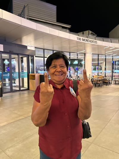 This media file is a vertical, full-body photograph of a person standing outside a modern building at night. The person, who appears to be of middle-aged or older, is smiling broadly and looking at the camera. They are wearing a maroon collared shirt and have their arms extended, with their index fingers pointing outwards, seemingly in a playful or defiant gesture. A black handbag is slung over their left shoulder.
Behind the person, the building features a sleek, metallic facade with geometric panels and large glass windows. The windows reveal interior lighting, suggesting the building is a commercial establishment, possibly a restaurant or retail space, with tables and chairs visible inside. The ground in front of the building is paved with light-colored tiles.
The text "100 MONTADITOS" is clearly visible in large, dark letters on a white panel on the building's exterior, indicating the name of the establishment. There are also some blue signs visible through the glass doors, though the text is largely illegible. The lighting suggests it is nighttime, and the scene appears to be well-lit. The overall mood is cheerful, with the subject's expression and gesture conveying a sense of fun and personality. The location context is Alcorcón, Spain.