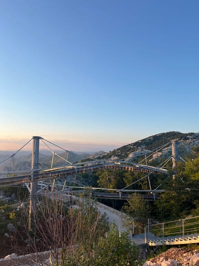 The image depicts a modern suspension bridge structure with elevated walkways winding through a mountainous, forested landscape. The bridge features tall, slender support columns and a network of cables supporting suspended pathways made of metal grating and wooden planks. The surrounding environment is characterized by rocky terrain, scattered trees, and rolling hills covered in dense vegetation. The sky is clear and blue, transitioning to a soft orange and pink hue near the horizon, suggesting either sunrise or sunset. In the foreground, a metal staircase with a railing leads down to a pathway. The scene appears to be a tourist attraction or recreational facility, given the constructed walkways designed for pedestrian access. The location is identified as being in Kuk, Montenegro, implying this is likely a viewpoint or a scenic trail in that region. There are no people visible in the image.